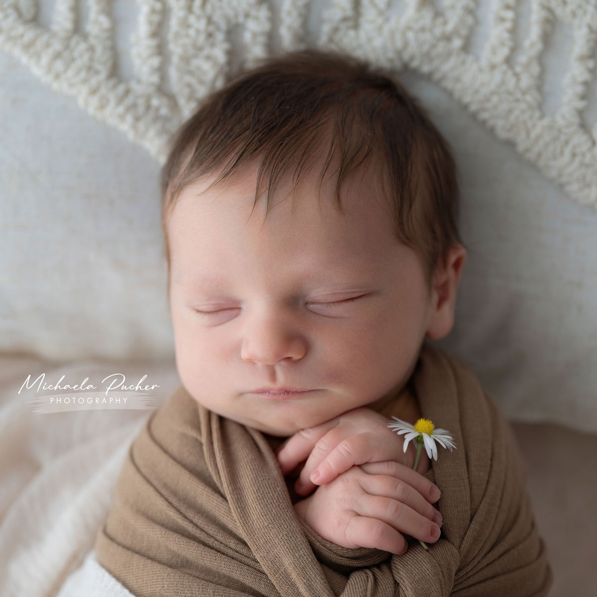Schlafendes Neugeborenes mit gefalteten Händen und einem kleinen Gänseblümchen, in braunes Tuch eingewickelt.